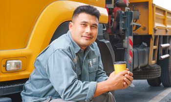 truck driver sitting on step of truck cab holding a coffeee in his hand